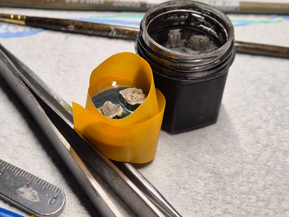 Close up of a base, filled with liquid water effect, and surrounded by messy, yellow tape. It's perched upon a metal tool, held in place by an old paint pot and another metal tool, all in the hopes, that it will be somewhat level in the end.