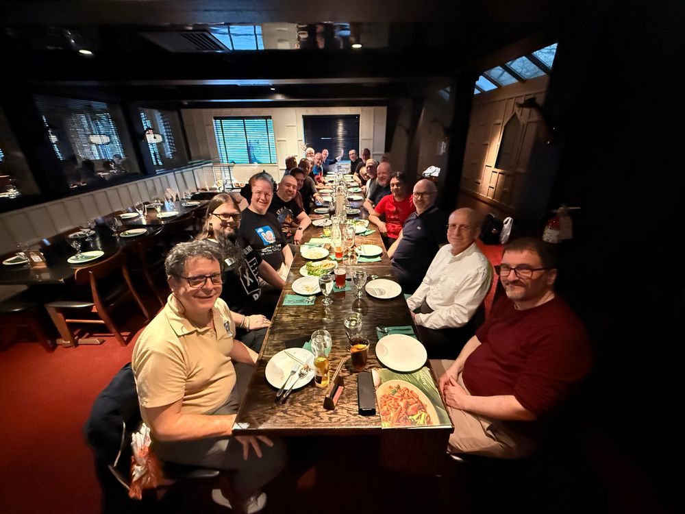 A group of people sit at a long table in a dimly lit restaurant. The table is set with plates, glasses, and various dishes. The restaurant has wooden paneling, large windows, and a cozy ambiance. The scene captures a social gathering of the Exchange MVP. Some bystanders have been removed via AI.