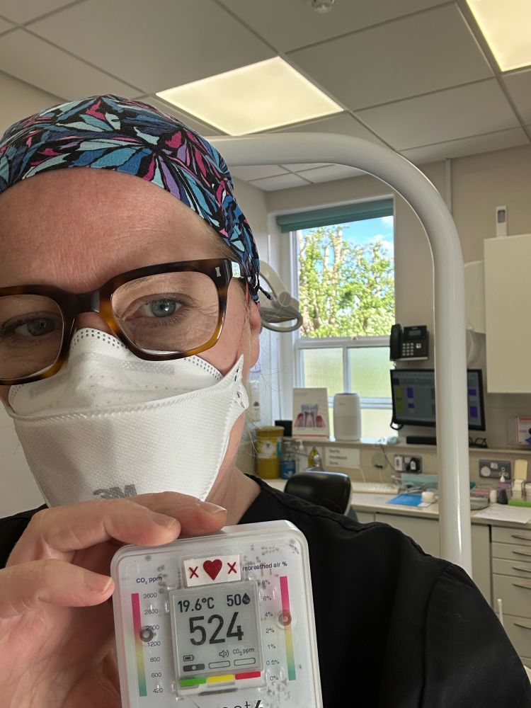 Dental hygienist wearing scrubs & hat & FFP3 respirator holds an Aranet4 C02 monitor reading 524ppm in a dental surgery with HEPA & FarUV & open windows.  