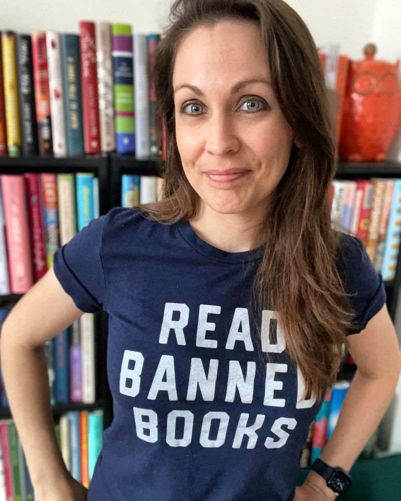 a photo of Kendra, a white woman with brunette hair, wearing a dark blue shirt with the words “read banned books on the front. Bookshelves can be seen behind her.