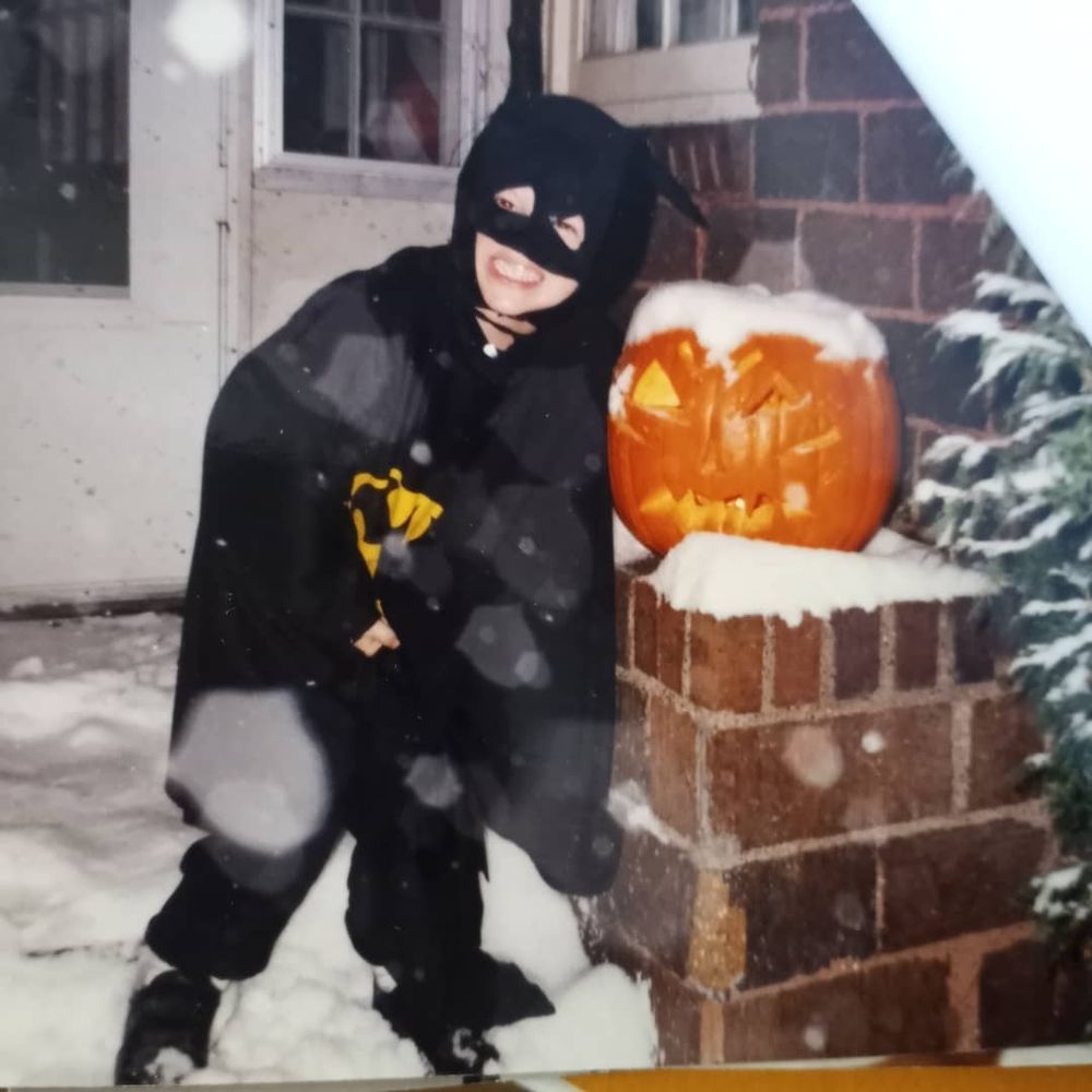 Me as a child in 1991 in a Batman costume on our front steps bending toward a big jack-o-lantern. Snow has covered our cedars and falling flecks are flaring out on the image.