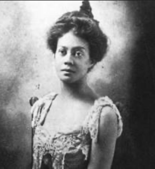A young black woman sits for a black-and-white portrait and a sleeveless dress with her hair in a bun on top of her head. 
