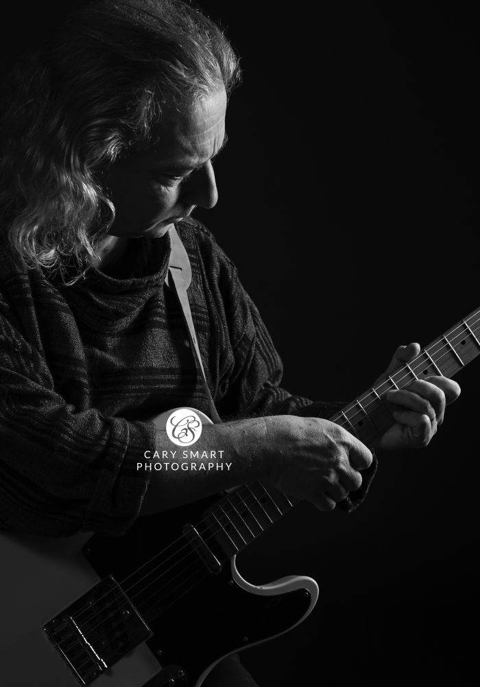Moody black and white image of an older male guitarist with long curly hair. The camera angle is diagonally down across the guitarist's chest from his right shoulder (camera left), looking down his face to where his hands are on the neck of the guitar as he's playing.  He's concentrating on his hands on the guitar.  The light is coming in from camera right and is just glancing off the guitarist, picking out the outline of the guitar body, the guitar strings, his fingers and the curve of his right hand, the guitar strap over his left shoulder, the profile of his face with a little light falling on his right check, and his curls over his right shoulder.