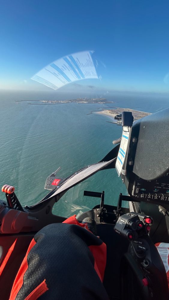 Blick aus dem Cockpit eines Hubschraubers über dem Meer: Instrumente, Steuerknüppel und meine Beine sind im Vordergrund zu sehen. Unter dem klaren blauen Himmel liegt Helgoland mit Hafenanlagen und Küstenlinien im Wasser.