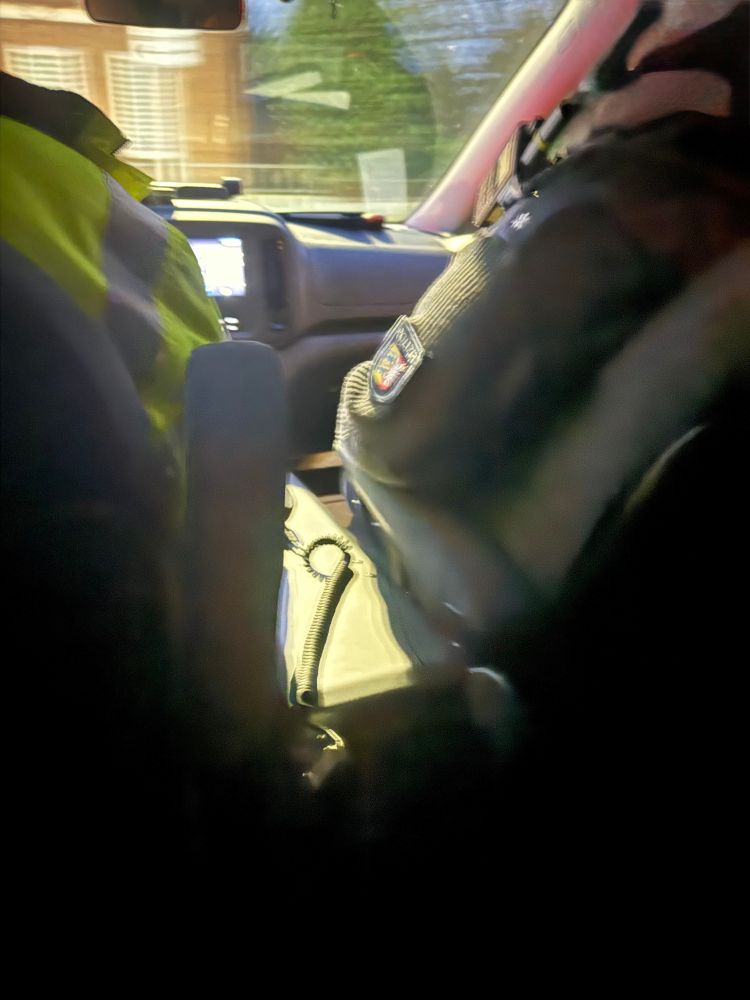 Blick aus dem Fonds in den Innenraum eines Einsatzfahrzeugs: Zwei uniformierte Polizist*innen sitzen vorne, zwischen ihnen ein Funkgerät und das Armaturenbrett. Durch die Frontscheibe sind unscharf Bäume und ein Gebäude zu sehen; die Bildränder sind dunkel und verschwommen.