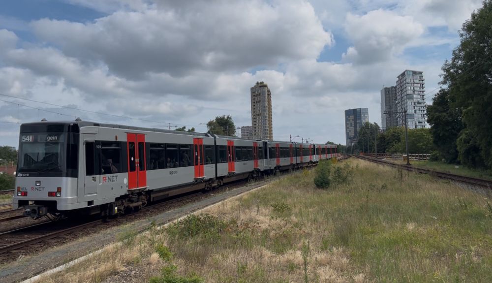 Silver and red Amsterdam metro M54 to Gien, on tracks to that go from the left side to the middle, the city centre can be seeing in the distance, there is another track on the other side, and there is some sparse grass in the area between the tracks before they merge