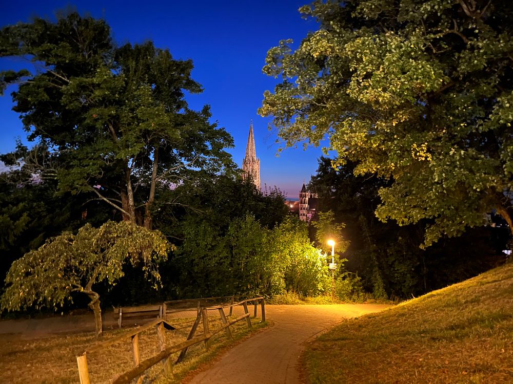 A path lit up kind of orange by a streetlamp, there’s the church and parts of the city visible behind the trees, and the path disappears behind a hill on the right side, there also a fence to one side and the path is coming from the cameras position, the sky is mostly a deep blue but it’s around sunset so near the horizon is more pink