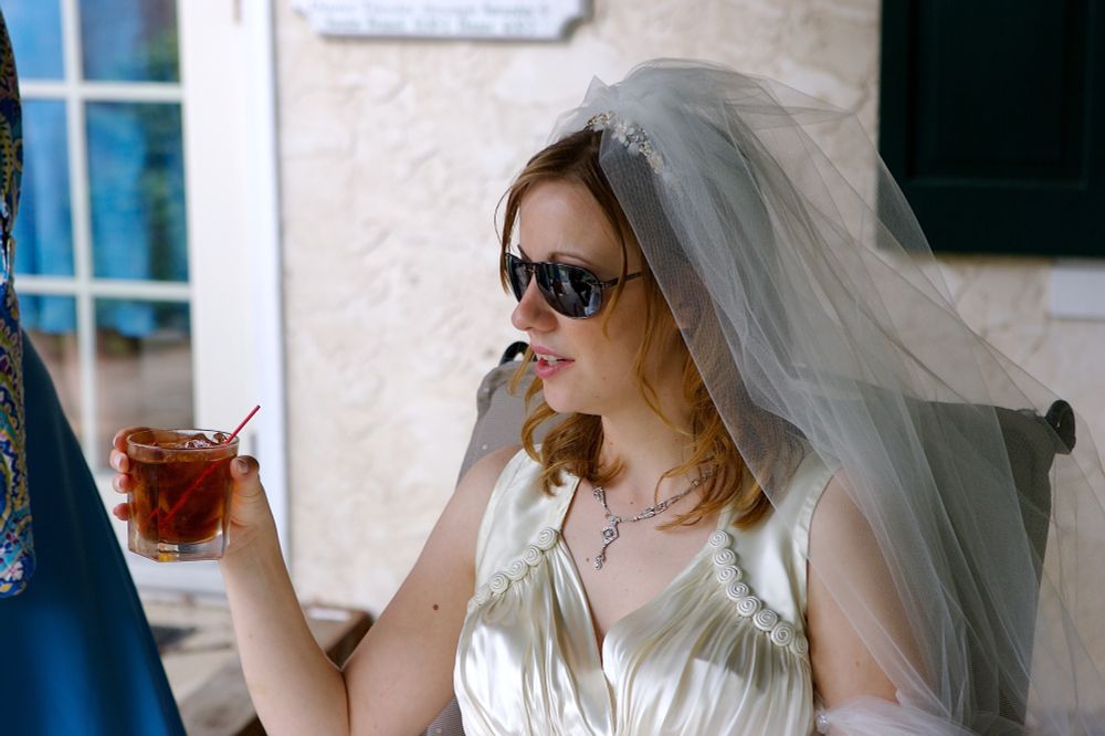 A beautiful woman in a satin wedding dress from the 40s wearing aviator sunglasses and holding a rocks glass filled with brown liquid while sitting in a chair. 