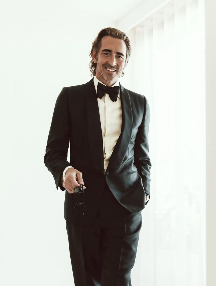 Lee Pace wearing a black suit, holding a pair of sunglasses in his hand. 