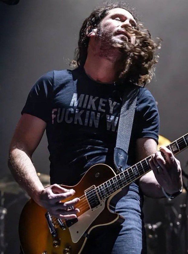 Photo of Ray Toro from My Chemical Romance with his head thrown back and eyes closed, looking majestic and beautiful as his hair is being tossed. He is playing his guitar and wearing a black shirt that says “Mikey Fuckin Way.” 