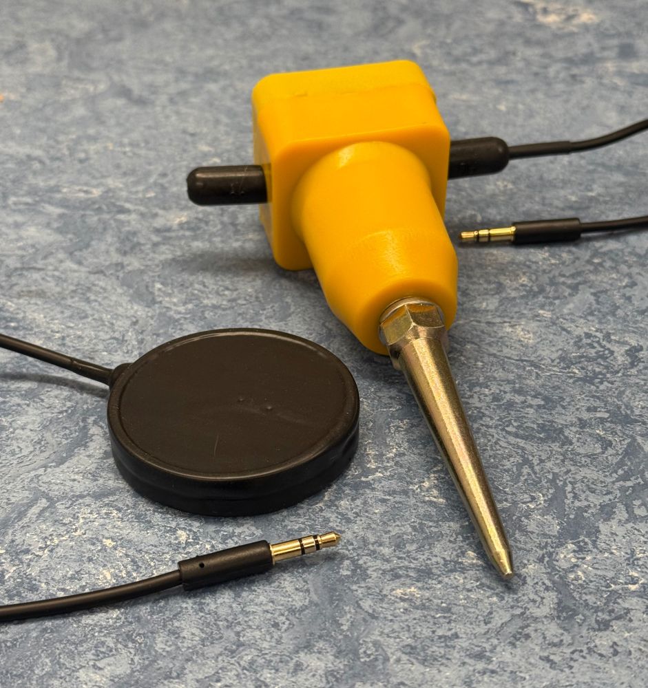 A photograph of a custom piezoelectric transducer sensor and a SM-24 geophone in a yellow commercial housing with a soil spike, sitting on a patterned workbench.