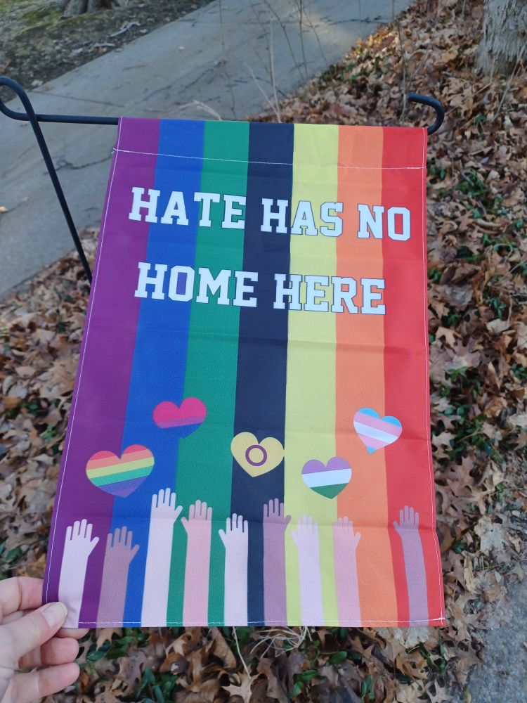 The flag states: Hate has no home here. The colors, left to right, purple, blue,green, black,yellow,orange, and red.  At the bottom of the flag, arms/hands of various shades are reaching towards five various colored hearts in the middle of the flag.