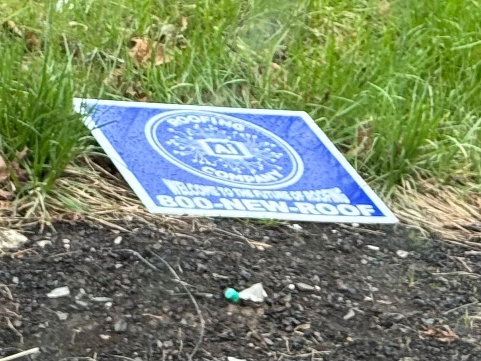 Belie sign with white lettering laying on the grass at the side of the road

The sign says has a large circular logo patterned after a circuit board. The logo has “AI” in the center and “Roofing Company”around the outside

Addition text says 
Welcome to the future OF ROOFING
800-NEW-ROOF