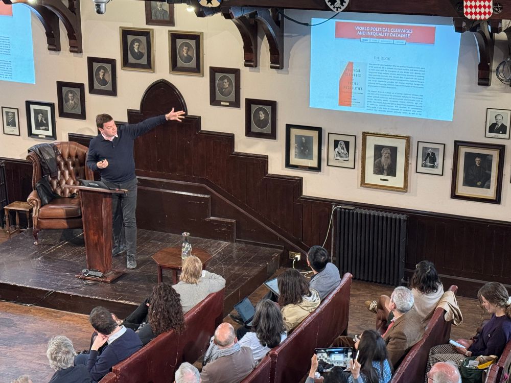 Thomas Piketty giving a lecture at the Cambridge Union. He is standing on the podium pointing to a slide showing his 2021 book Political Cleavages and Social Inequalities.