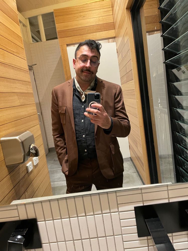 A man in his mid-30s with dark hair and glasses wearing a brown suit, black shirt and neckerchief taking a selfie in a bathroom at his Aunt’s second wedding 