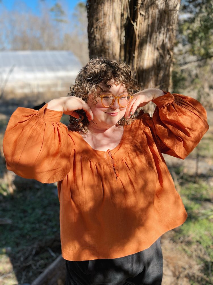 I’m posing in front of a tree in dappled sunlight. Wearing an orange gathered blouse with large gathered sleeves. There are 4 buttons down a front half-placket. 