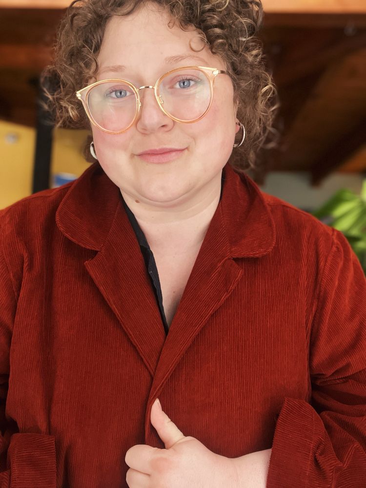 A white woman with blonde curly hair up in a bun and orange glasses in a rust orange-red corduroy chore coat with no buttons 
