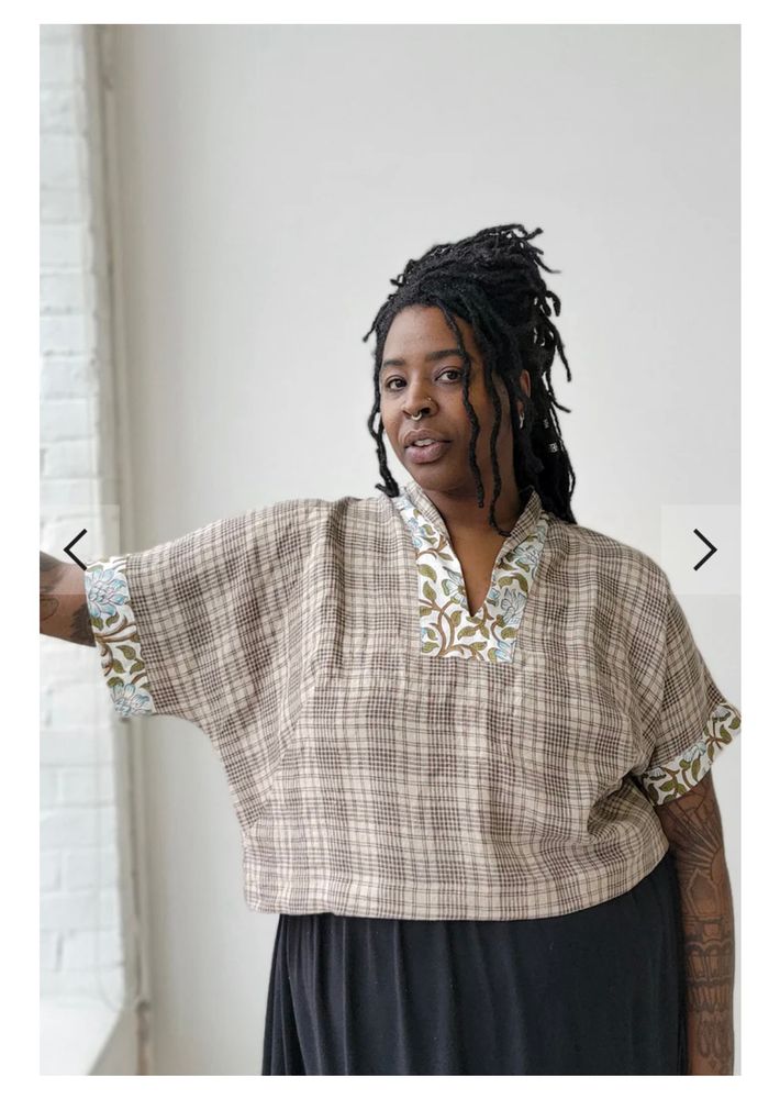 A model with braids pulled back in a half-up do is wearing a linen, dolman sleeve blouse with a contrast facing and contrast cuffs and a shawl collar. The main fabric is a cream and brown plaid and the contrast fabric is a white, green and blue floral pattern. 