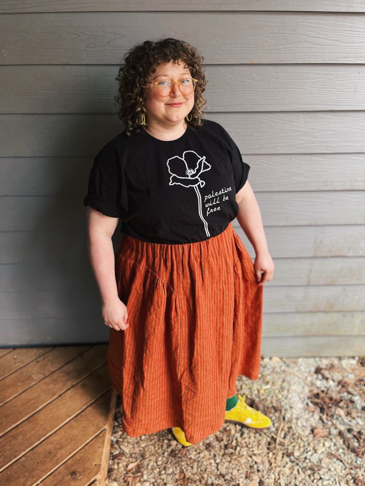 Woman with blonde curly hair (me!) standing showing off her newly sewed gathered elastic waist skirt in orange and tan stripes. Wearing yellow sneaker, green socks and a shirt that says “Palestine will be free” 