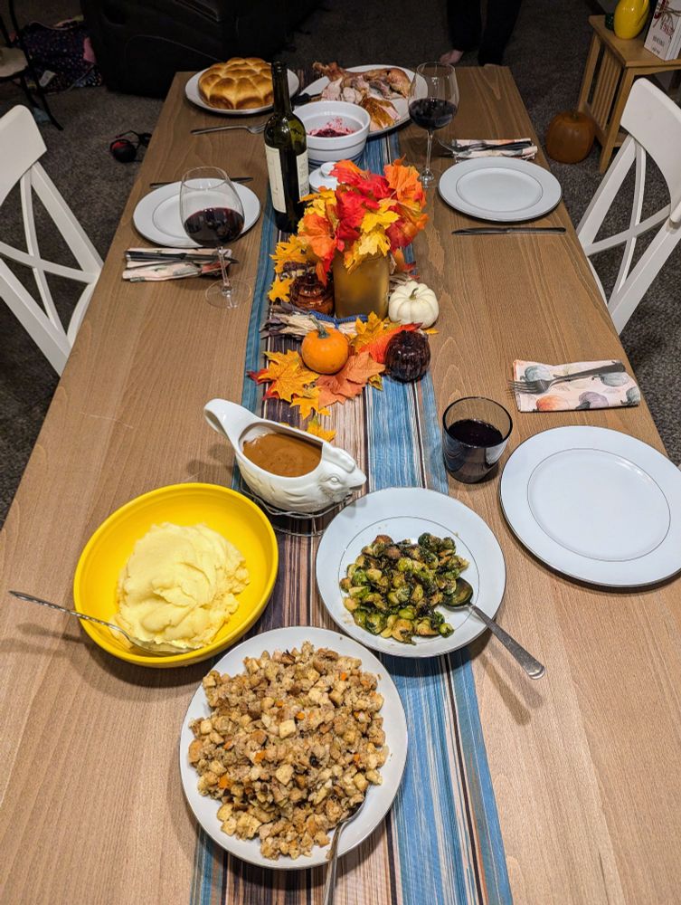 Table set with various thanksgiving dishes and a centerpiece made of fake leaves and real gourds.