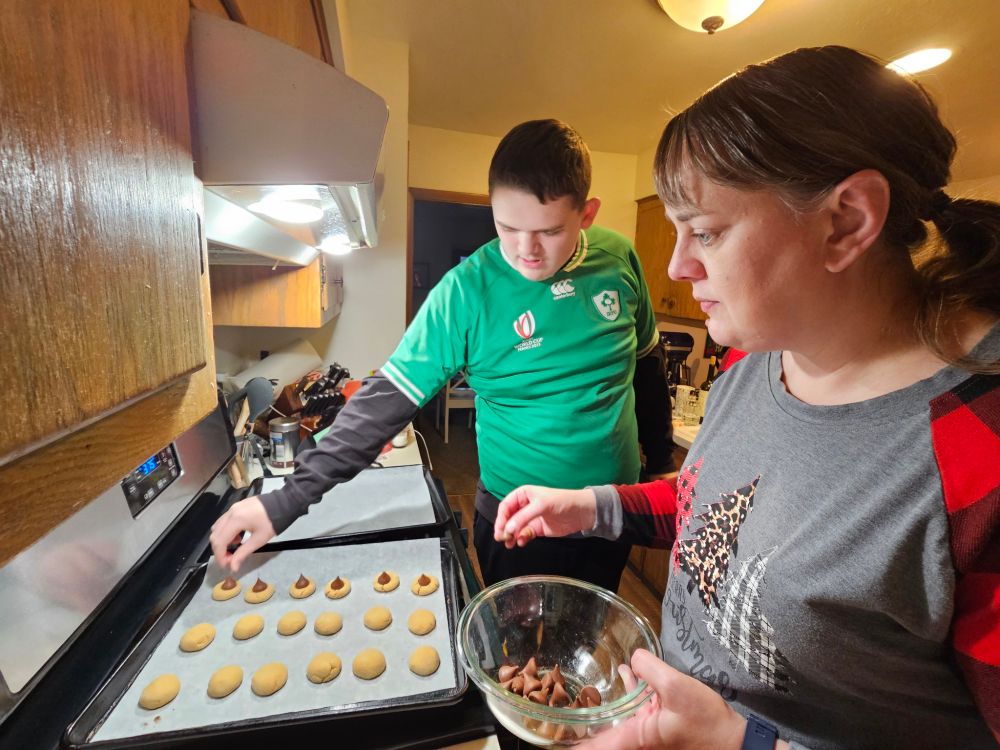 My giant child (he's 4 inches taller than me) in the green shirt helping place Hershey Kisses on peanut butter cookies. I'm in a gray shirt with plaid Christmas trees and am holding the bowl of Hershey Kisses. 