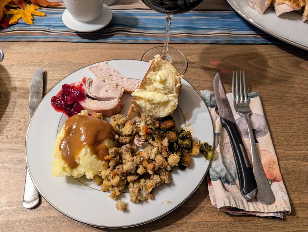 Full plate of roll with butter, brussel sprouts, stuffing, mashed potatoes with gravy, turkey and cranberry sauce. 