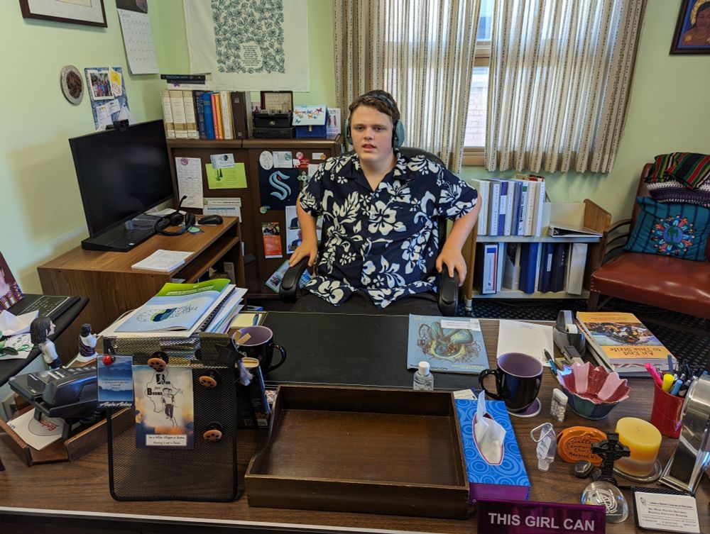 Teenage boy wearing a blue Hawaiian shirt and wearing light blue headphones sitting at a semi -cluttered desk in an office, with a monitor to his left and a window behind him with curtains.