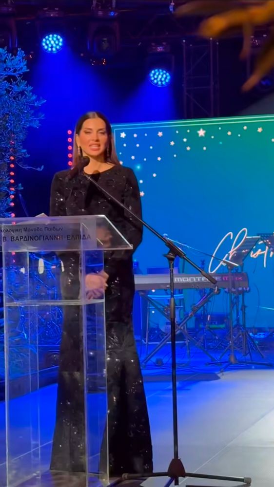 Apresentadora Stamatina de vestido preto de lantejoulas discursando num jantar de caridade.