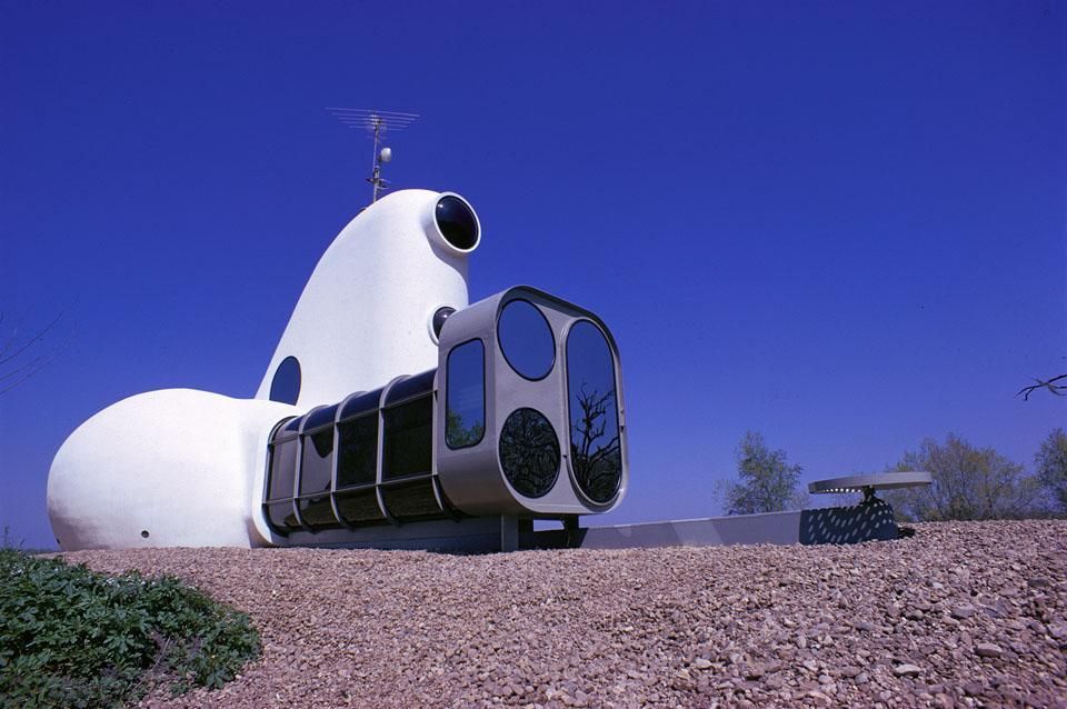 Futuristic blobby shaped house with rounded windows and a geometric glass entryway, sitting on mulched ground under a blue sky.