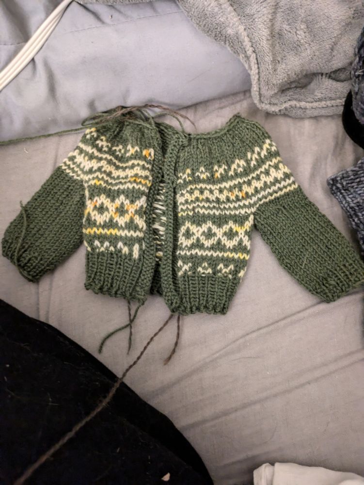 A photo of a doll sized army green and butter yellow colorwork cardigan. The body is dark green with a yellow colorwork chart that normally would only be in the yoke. The cardigan has been cut open and you can see many ends of yarn.