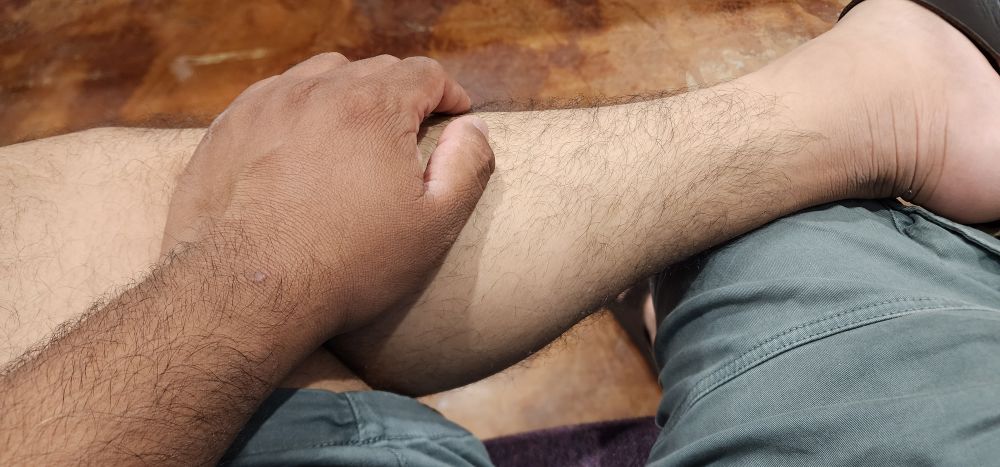 A photo of a hand resting on a leg as a man in shorts sits with one leg crossed over the other. His arm is darker than his leg