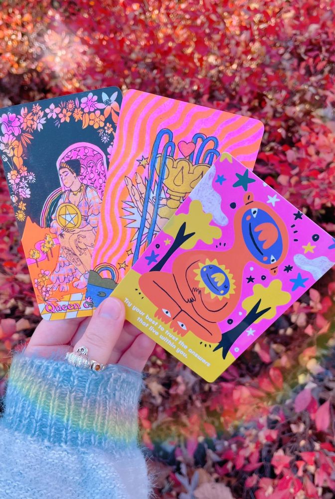my hand (white and hobbit-sized) holding three tarot and oracle cards in front of blueberry bushes-turned-autumn red. the cards pictured from left to right are: the queen of pentacles, the ace of cups (both from the queer tarot), and an oracle card from the 'gentle reminders' deck by barry lee. the oracle card features a stylized and smiling muppet-like monster with a big nose, holding a smaller, similar-shaped creature over their heart-space. the card reads, "try your best to trust the answers that live within you." a silver pumpkin-shaped ring is on my thumb. the photo has been edited to add a rainbow lens flare and saturation, especially in favor of the warmer hues.