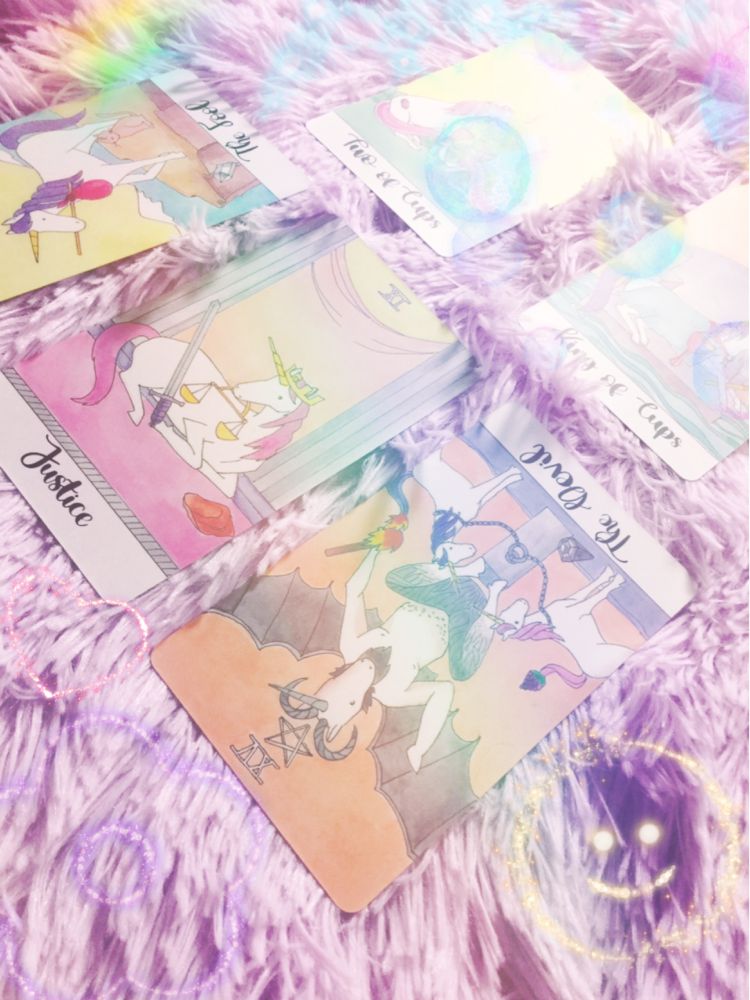 a five-card spread from the crystal unicorn tarot. the cards pictured are the two of cups, king of cups, the fool reversed, justice, and the devil reversed. the cards are placed on a fluffy pastel purple blanket. the photo's been edited to add rainbows, sparkles, and bubbles, adding a whimsical aesthetic.