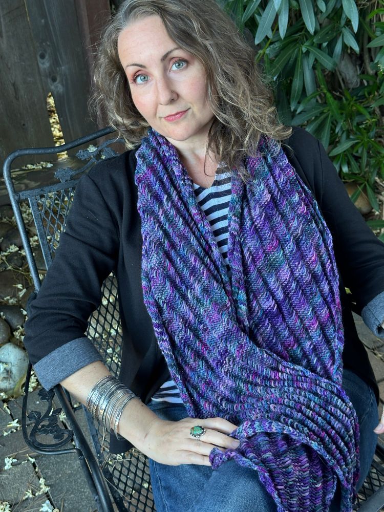 Woman seated wearing a handknit variegated purple scarf 