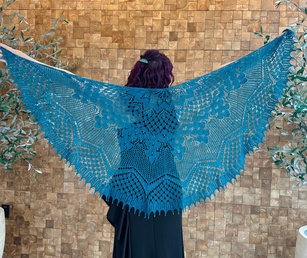 Woman facing away holding up an elaborate beaded lace shawl knit in teal yarn