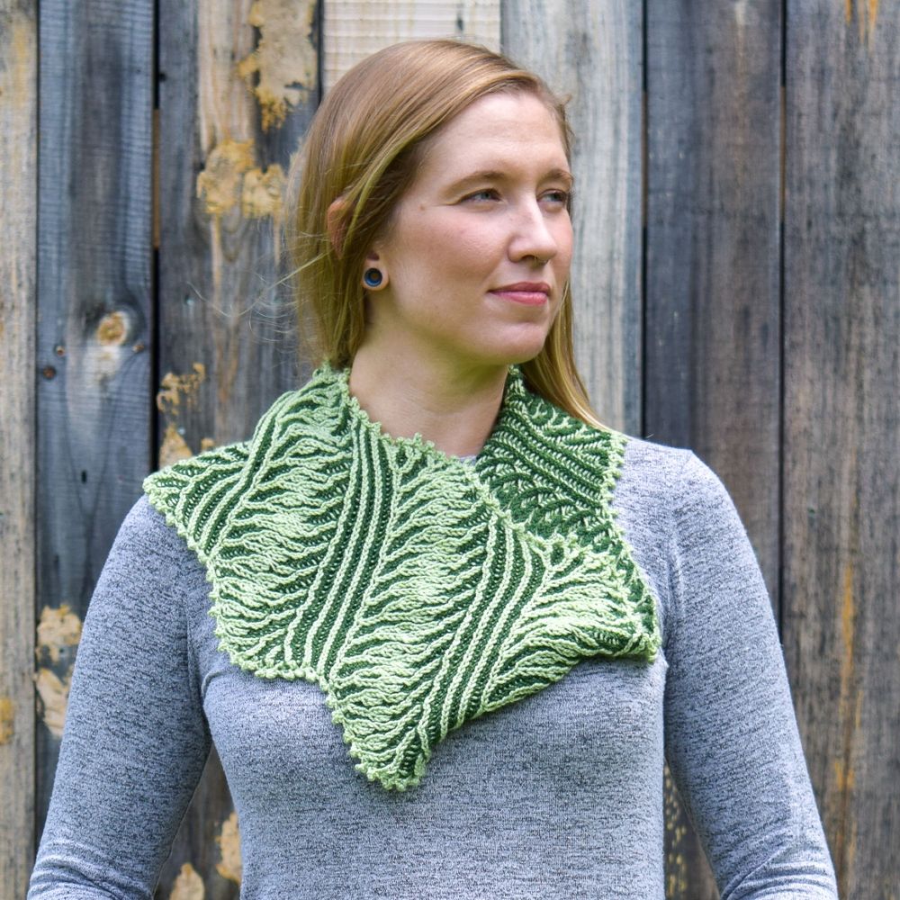 A woman is wearing a hand knit brioche stitch cowl in a swirly pattern in two shades of green.