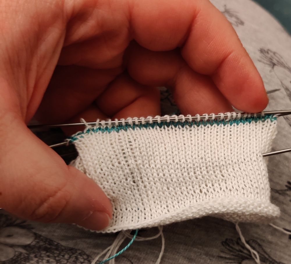 A hand is holding a knitted white tube with very fine metallic needles.