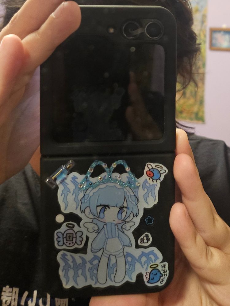 capinha customizada de samsung flip 5 com um adesivo com uma pessoa vestida na moda japonesa "Tenshi Kaiwai" no meio e adesivos menores de tamagotchis anjos aos lado, assim como duas asinhas. a capinha é preta e os adesivos são quase todos brancos ou azuis.