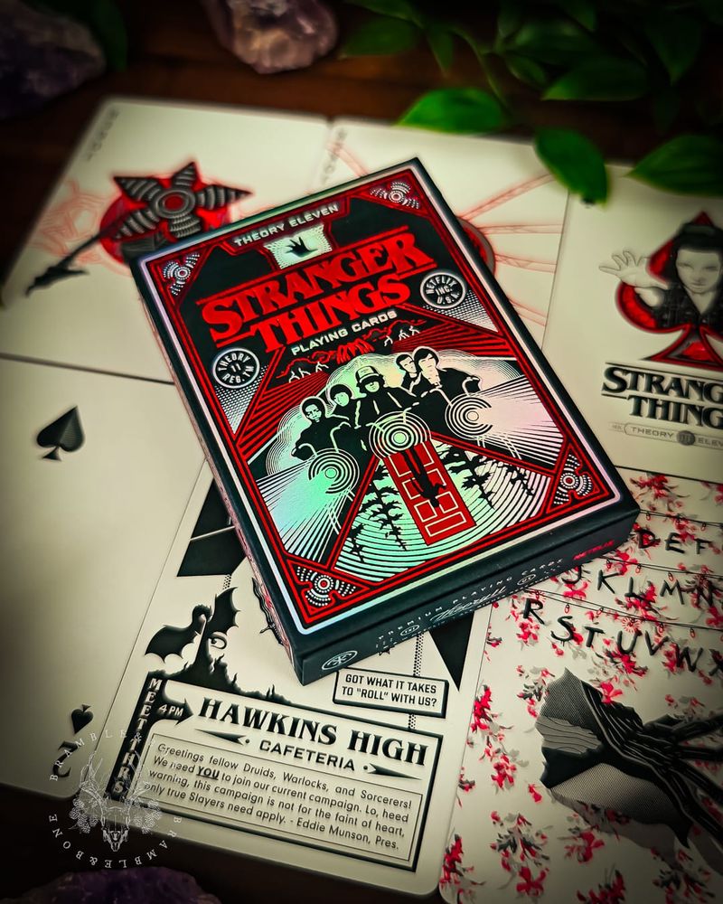 Hand-illustrated Stranger Things playing cards spread on dark wood, showing custom face cards with intricate red and black artwork depicting series characters. The Jack, Queen, and King of clubs feature Will, Jonathan, and another character in ornate traditional playing card styling with cameras, axes, and iconic show elements woven into detailed patterns.