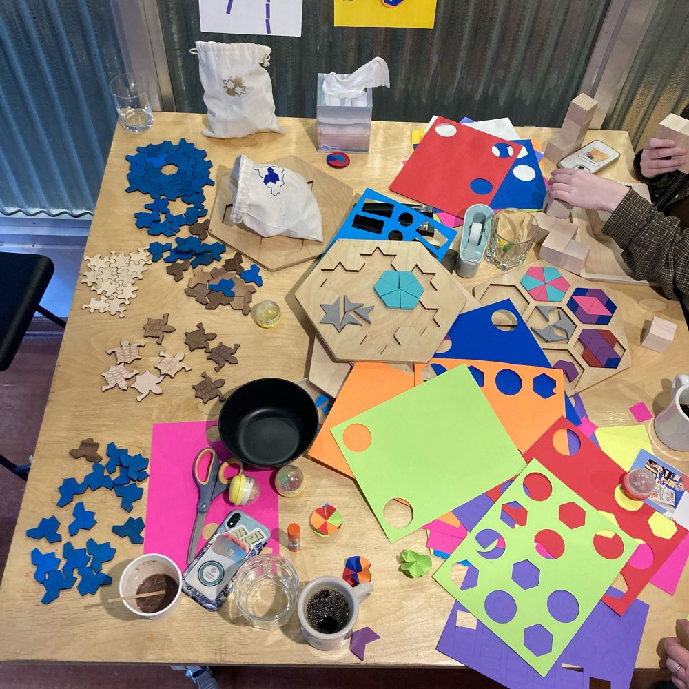 Looking down on a large square tabletop covered with a diverse collection of colorful shapes and tiles and at least two cups of coffee