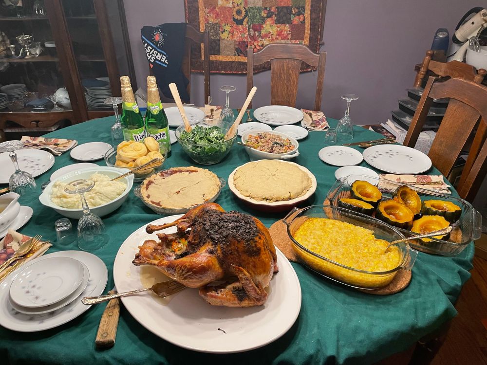 Dinner table with dark green cloth, presenting Thanksgiving feast of turkey, corn casserole, mashed potatoes, acorn squash, rolls, salad, and two pies. 