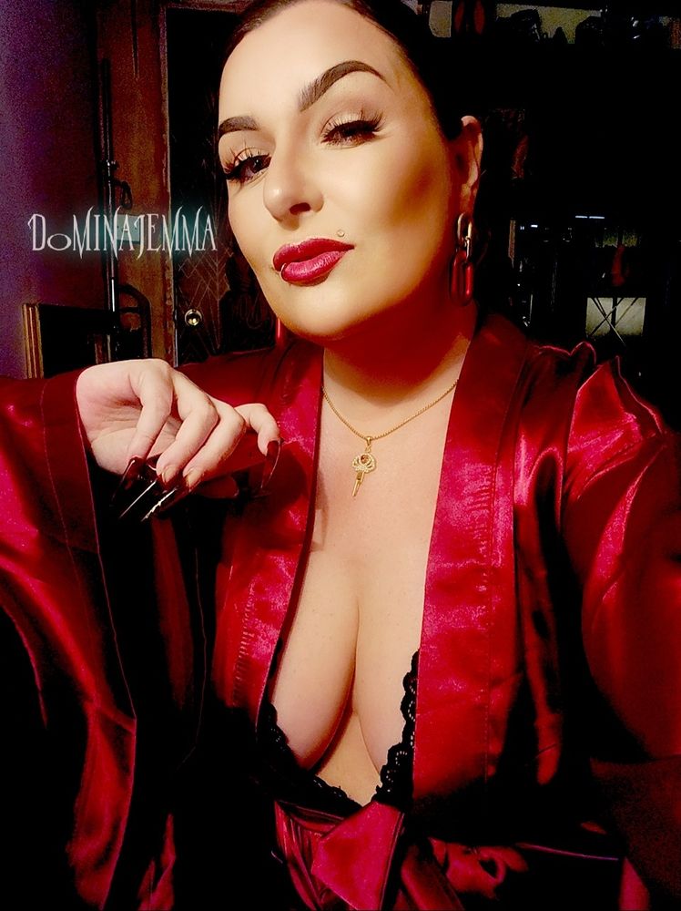 Domina Jemma wears a burgundy satin kimono robe with a peak of black lace revealing her cleavage. She wears a gold necklace that has a crystal crown chastity key pendant, and red and hold chain link earrings. 