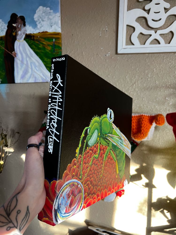 A side view of a painting of a gold-green sweat bee on a red flower. The bee and flower sit on a black background, on the side edge of the canvas is the artist’s signature and script as follows: “2025. K. Mitchell (2485), Ottawa Nation of Oklahoma” 