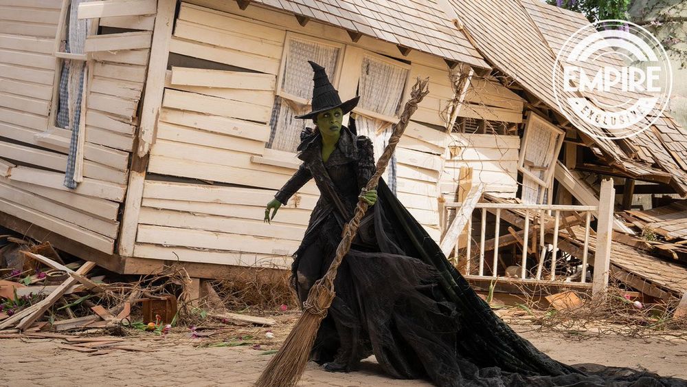 Elphaba diante da mesma casa de madeira branca destroçada. Ela parece estar andando, segurando a vassoura em uma mão e olhando para fora do enquadramento, usando o chapéu pontudo.