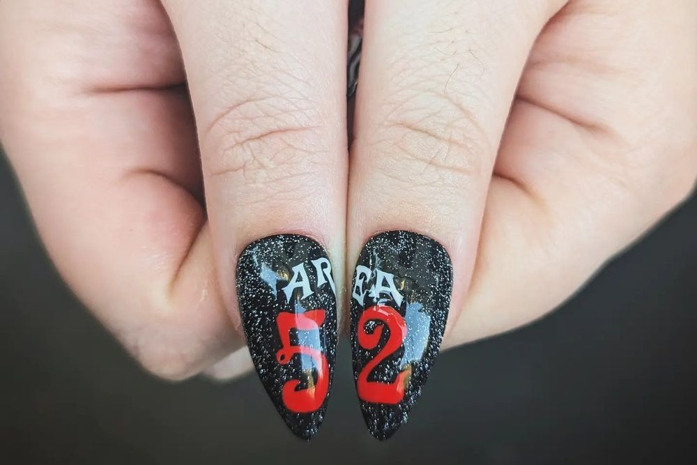 A pair of thumbs put together to showcase the press on nails applied on them. The logo of the Area 52 tour is complete when the two nails are next to each other. It reads AREA in white letters, then 52 in big red letters, on a black background with reflective glitter.