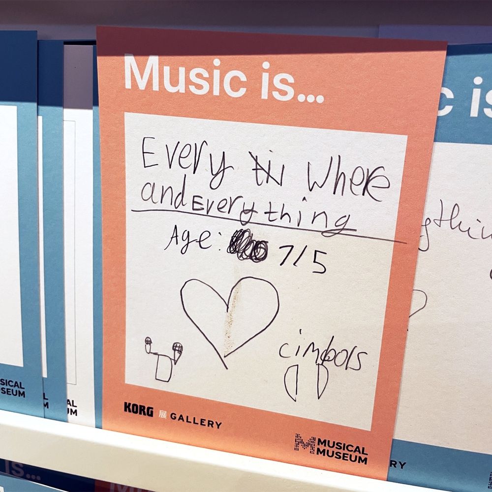 A note headed "Music is..." Underneath, someone has written in black pen: "Everywhere and everything. Age 7/5". Below is a hand-drawn heart, along with the word "cimbols" and two drawn cymbals.