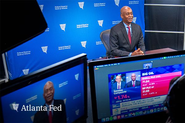 Atlanta Fed president and CEO Raphael Bostic sits at a table in front of a Federal Reserve Bank of Atlanta backdrop in a studio talking to a television monitor.