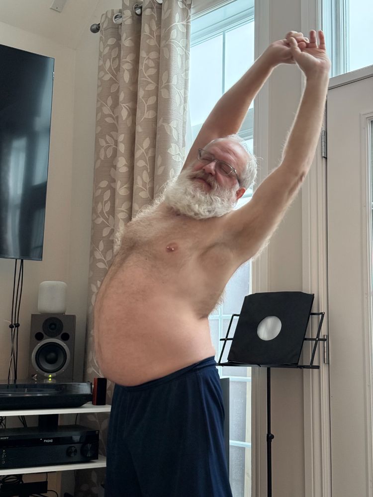 White haired, white bearded man shirtless wearing blue shorts, stretching with his arms upward, standing in front of a tall window and various stereo equipment.