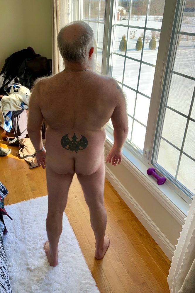 Nude man shown from behind standing with one foot on a white rug, one foot on a hardwood floor, looking out a window at a snow-covered backyard.