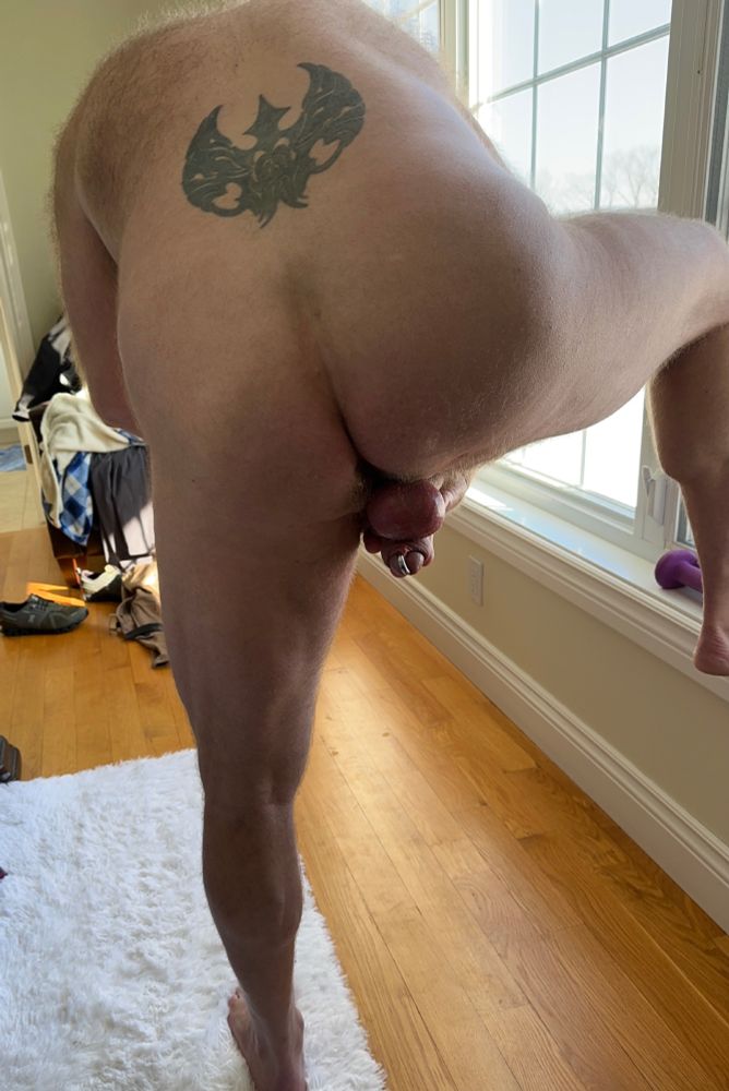 Nude man shown from behind standing with one foot on the floor, the other foot on the windowsill, bending over and pushing his cock and balls back towards his butt crack.